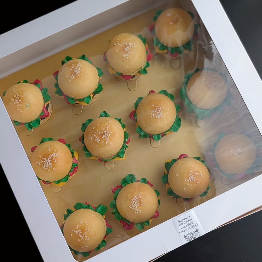 Box of 12 novelty burger cupcakes on a gold board — each built from two cupcake "buns" with a chocolate cake "patty," hand-piped green lettuce, yellow cheese and red tomato buttercream, and a sesame seed topping