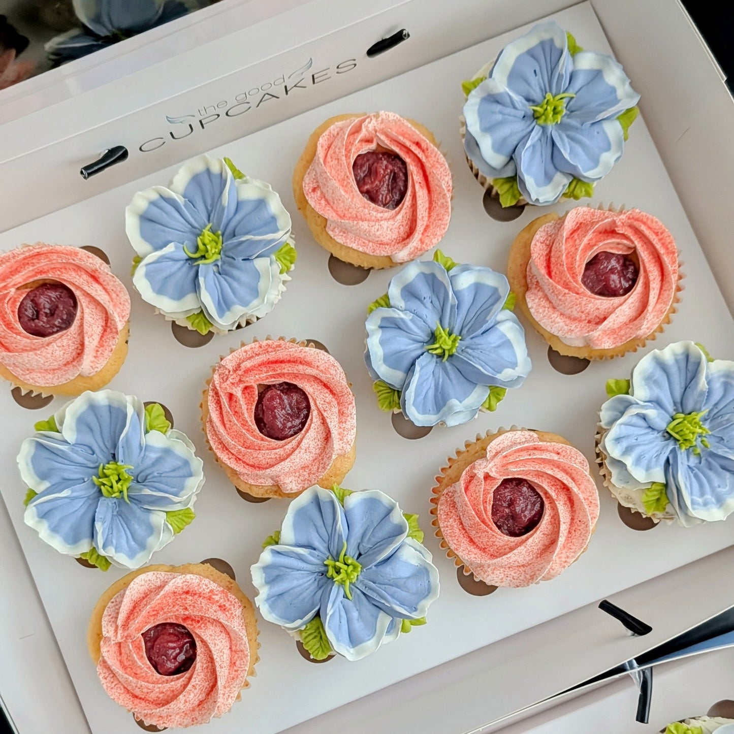 Box of 12 cupcakes — six pink strawberry rosette cupcakes with a cherry centre and six hand-piped blue five-petal flower cupcakes with a green centre, in a white gift box labelled The Good Cupcakes