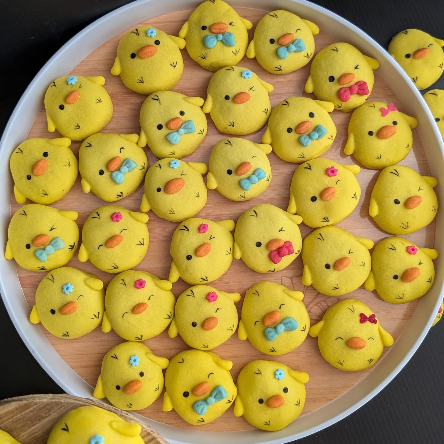 A round tray filled with small yellow chick-shaped baked goods decorated with colorful eyes, beaks, and bow ties, set against a dark background.