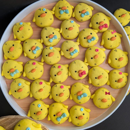 A round tray filled with small yellow chick-shaped baked goods decorated with colorful eyes, beaks, and bow ties, set against a dark background.