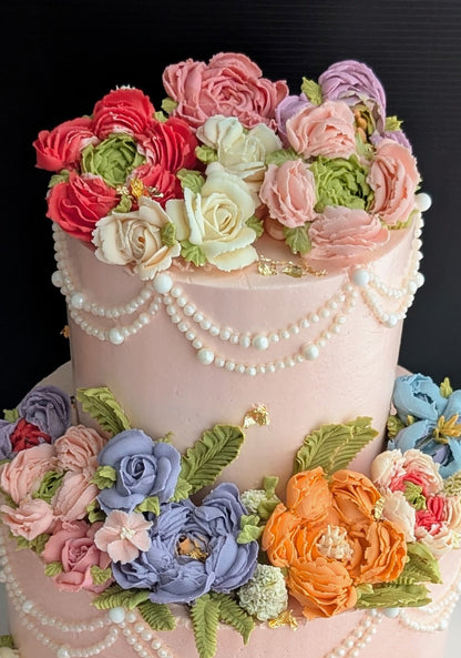 This is a two-tiered pink cake with cascading pearl draping and an abundance of hand-piped multicolour buttercream flowers (roses, peonies, ranunculus in pink, purple, blue, orange, and white).