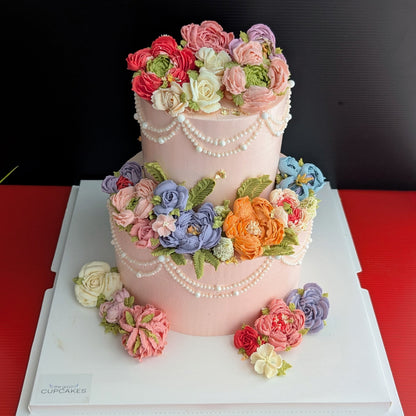 This is a two-tiered pink cake with cascading pearl draping and an abundance of hand-piped multicolour buttercream flowers (roses, peonies, ranunculus in pink, purple, blue, orange, and white).
