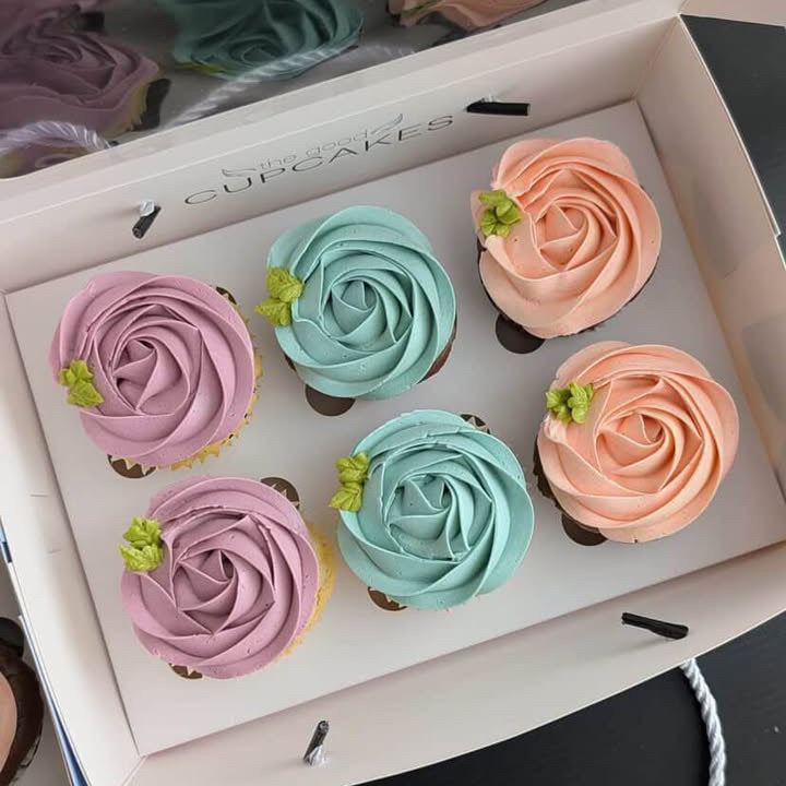 Six rosette flower cupcakes by The Good Cupcakes Company, with rosette-shaped frosting in pastel pink, mint green, and peach, presented in a white bakery box with a clear lid.
