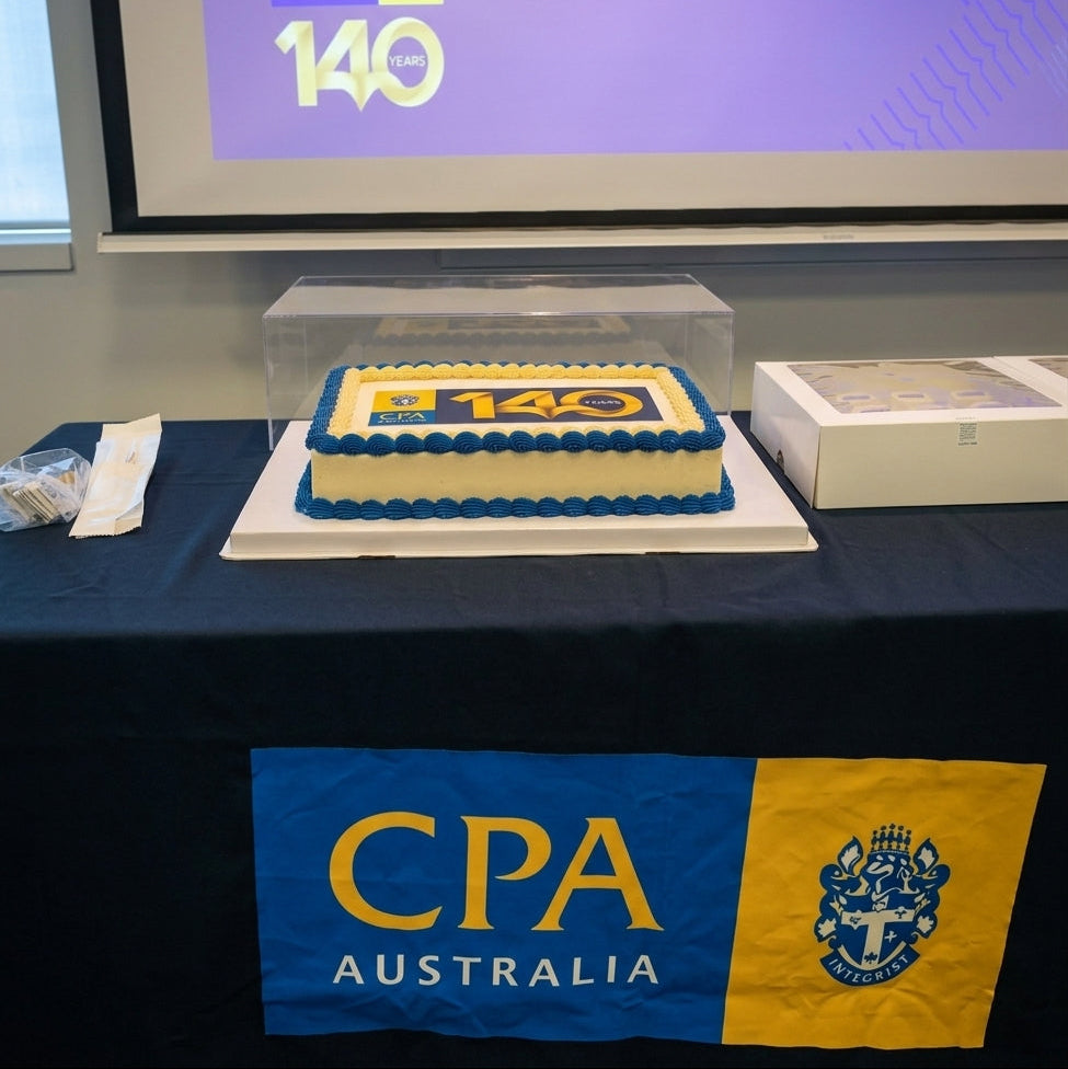 Blue and yellow CPA Australia 140 Years logo cake displayed beneath the anniversary presentation screen, with boxed cupcakes alongside.