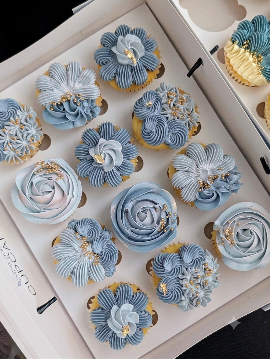 Box of 12 gender reveal cupcakes with hand-piped blue floral buttercream in baby blues, chrysanthemum and petal fan designs with gold leaf and pearl sprinkles, The Good Cupcakes Singapore