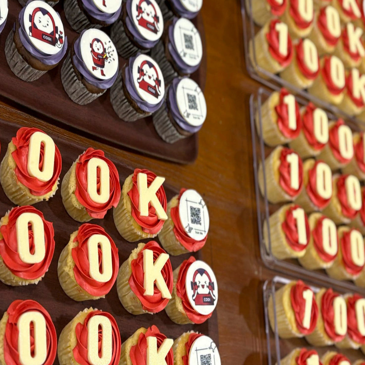 Red and purple cupcakes decorated with 100K lettering, CODI character toppers and QR codes, created for IMDA’s celebration of reaching 100,000 LinkedIn followers.