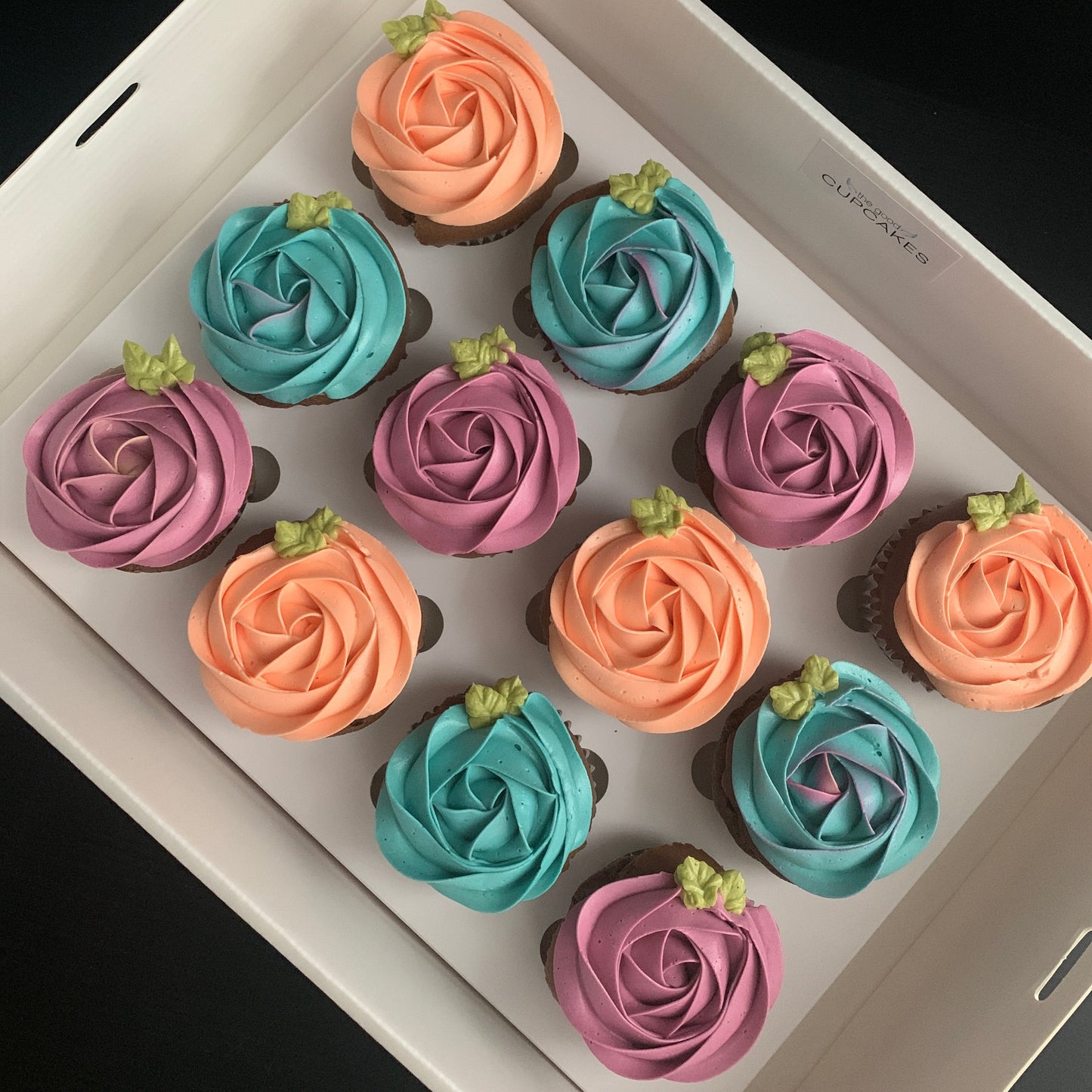 Box of 12 beautiful low calorie cupcakes by The Good Cupcakes Company, each in a rosette floral design in various colours.