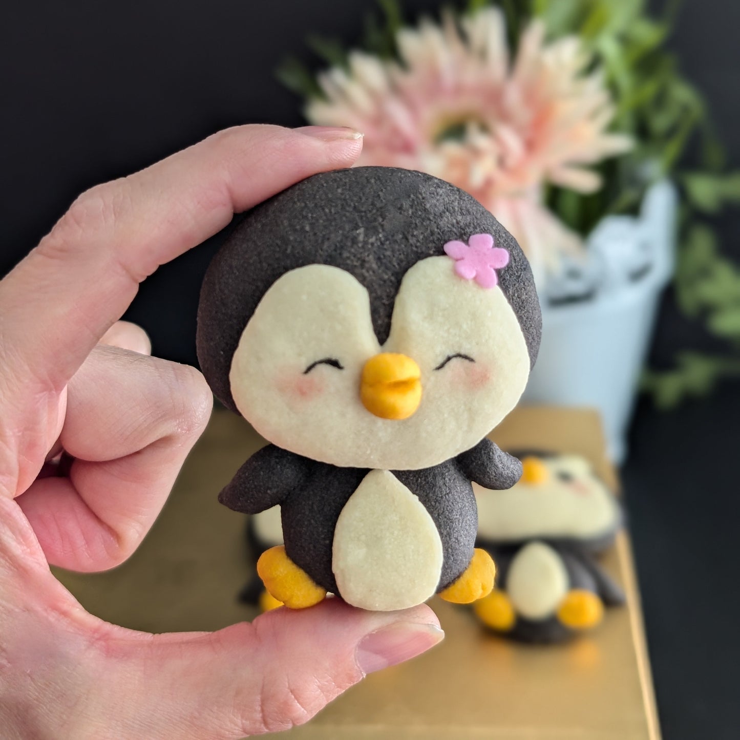 Handmade penguin-shaped shortbread cookie held in a hand — black and cream cookie with hand-sculpted yellow beak and feet, rosy cheeks, and a pink flower detail. Made to order by The Good Cupcakes, Singapore.
