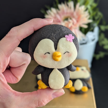 Handmade penguin-shaped shortbread cookie held in a hand — black and cream cookie with hand-sculpted yellow beak and feet, rosy cheeks, and a pink flower detail. Made to order by The Good Cupcakes, Singapore.
