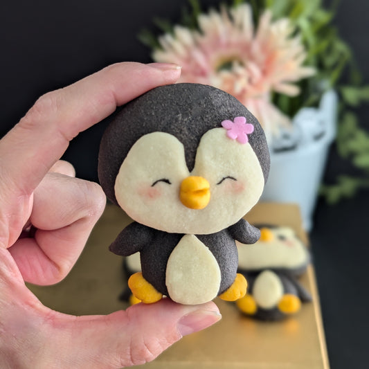 Handmade penguin-shaped shortbread cookie held in a hand — black and cream cookie with hand-sculpted yellow beak and feet, rosy cheeks, and a pink flower detail. Made to order by The Good Cupcakes, Singapore.

