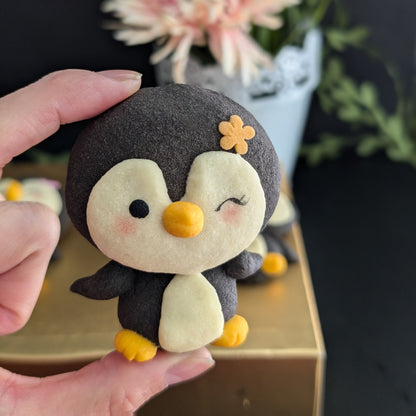 A handmade felt penguin plush toy being held by a hand. The penguin has a dark charcoal gray body, a cream-colored face and belly, a yellow beak and feet, and a small yellow flower on its head.