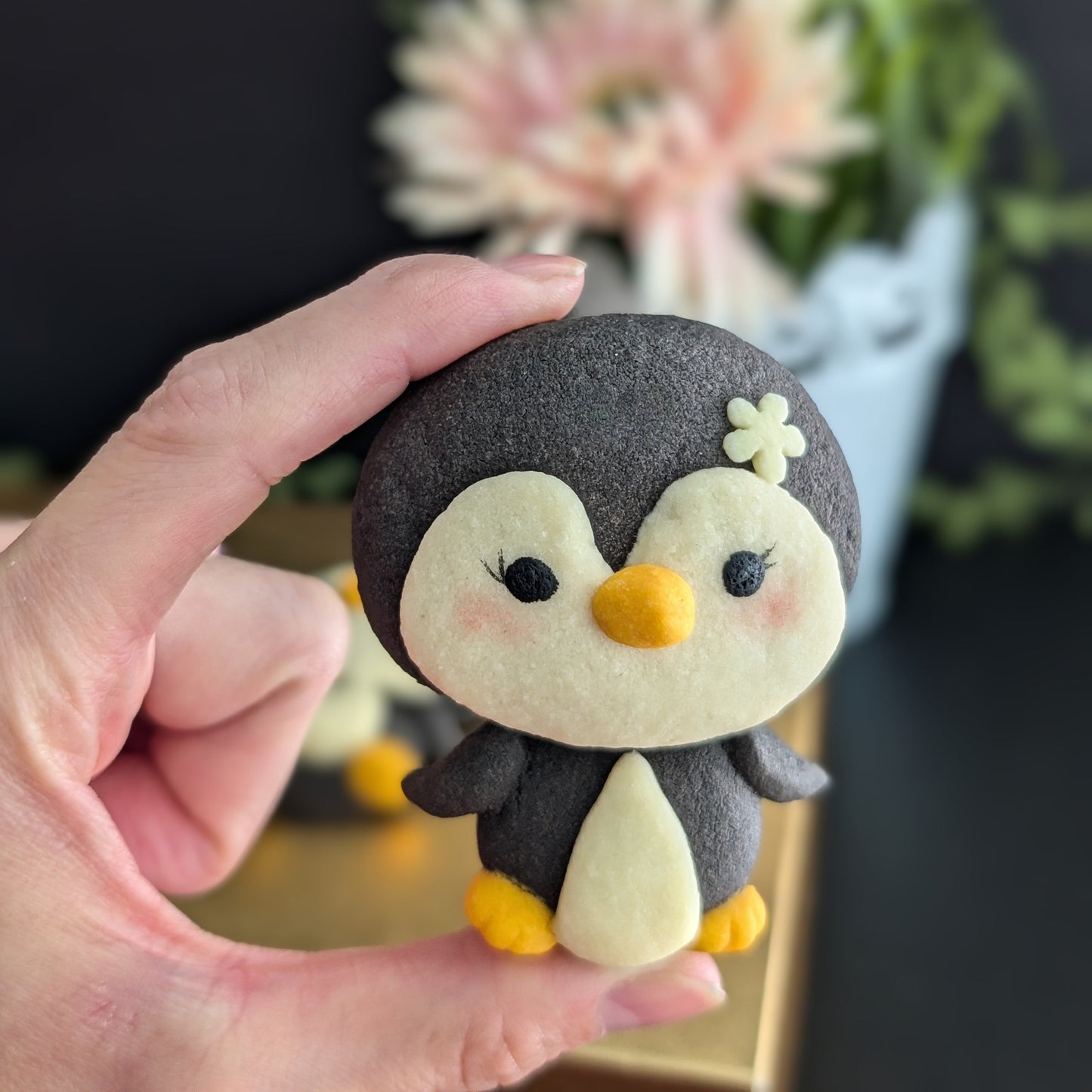 A hand holds a small felt penguin toy with a dark charcoal gray body, cream-colored face and belly, yellow beak and feet, and a small cream flower on its head. The background is a soft-focus indoor setting with a dark wall and blurred flowers.
