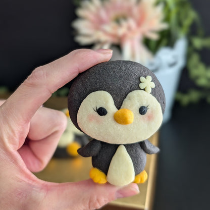 A hand holds a small felt penguin toy with a dark charcoal gray body, cream-colored face and belly, yellow beak and feet, and a small cream flower on its head. The background is a soft-focus indoor setting with a dark wall and blurred flowers.
