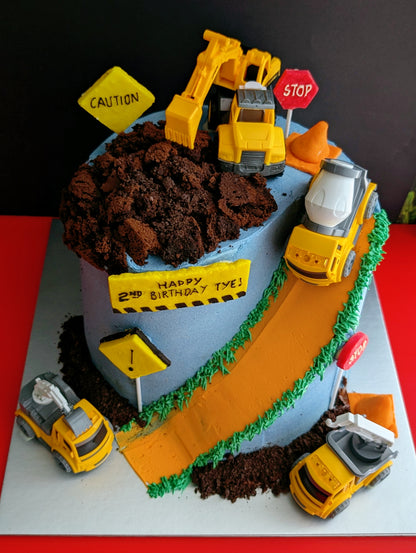 Construction site birthday cake with toy trucks, excavator, cement mixer, chocolate dirt, orange road and green grass buttercream details, low-sugar cake Singapore