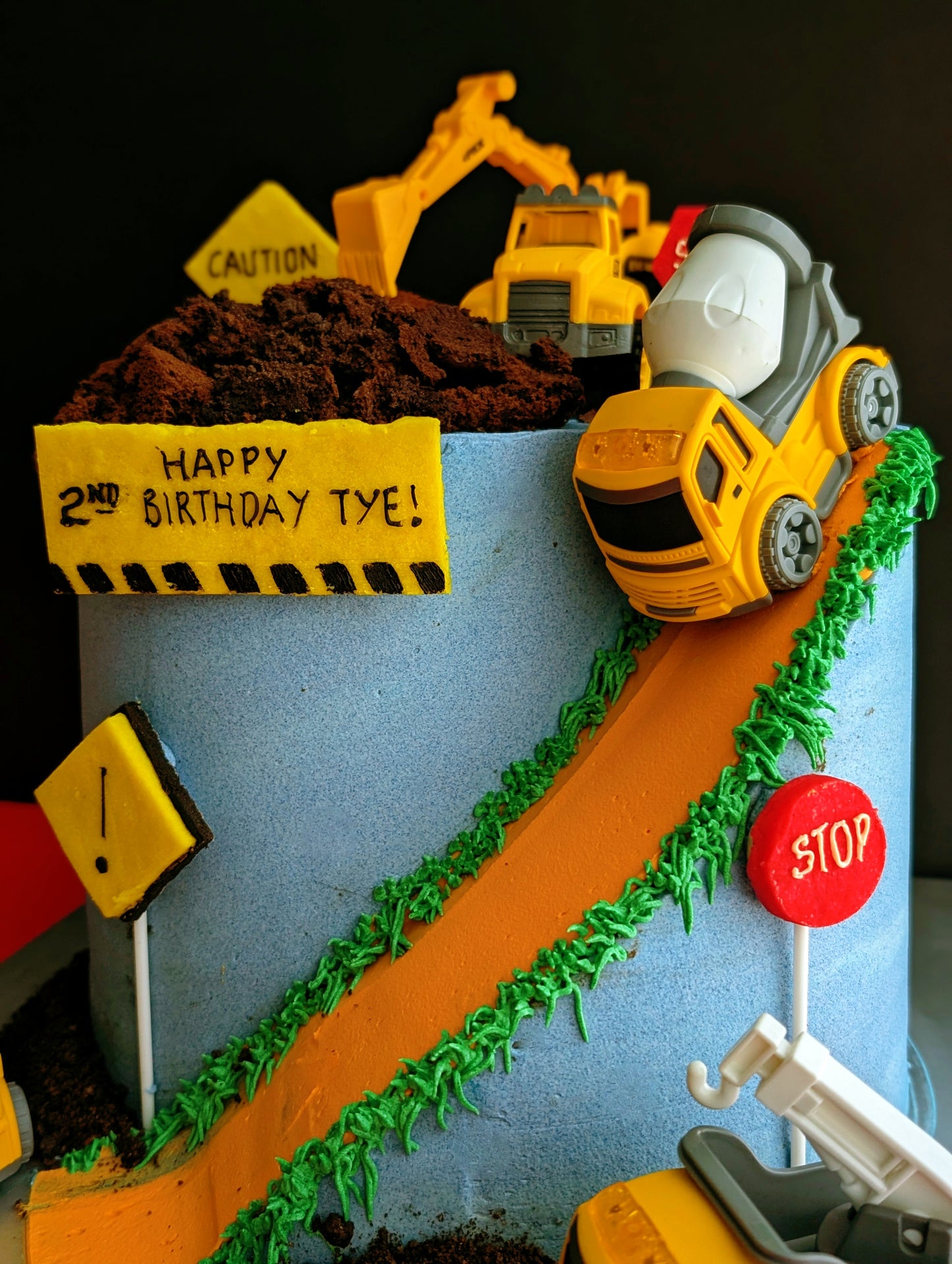 Construction site birthday cake with toy trucks, excavator, cement mixer, chocolate dirt, orange road and green grass buttercream details, low-sugar cake Singapore