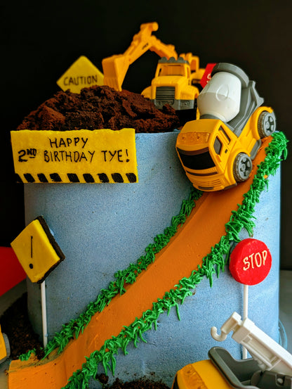 Construction site birthday cake with toy trucks, excavator, cement mixer, chocolate dirt, orange road and green grass buttercream details, low-sugar cake Singapore