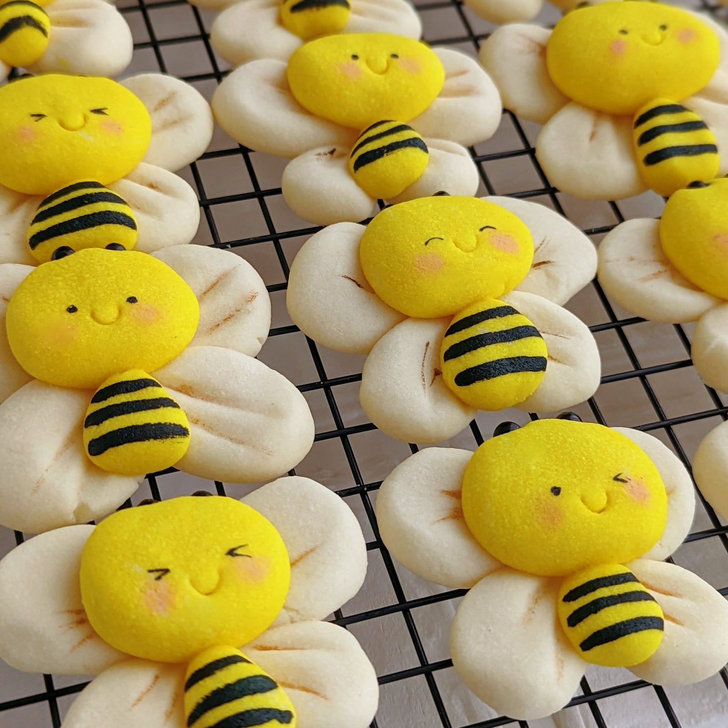 A close-up of several cute baked cookies shaped like smiling yellow bees with black stripes and cream wings, arranged on a black wire cooling rack.