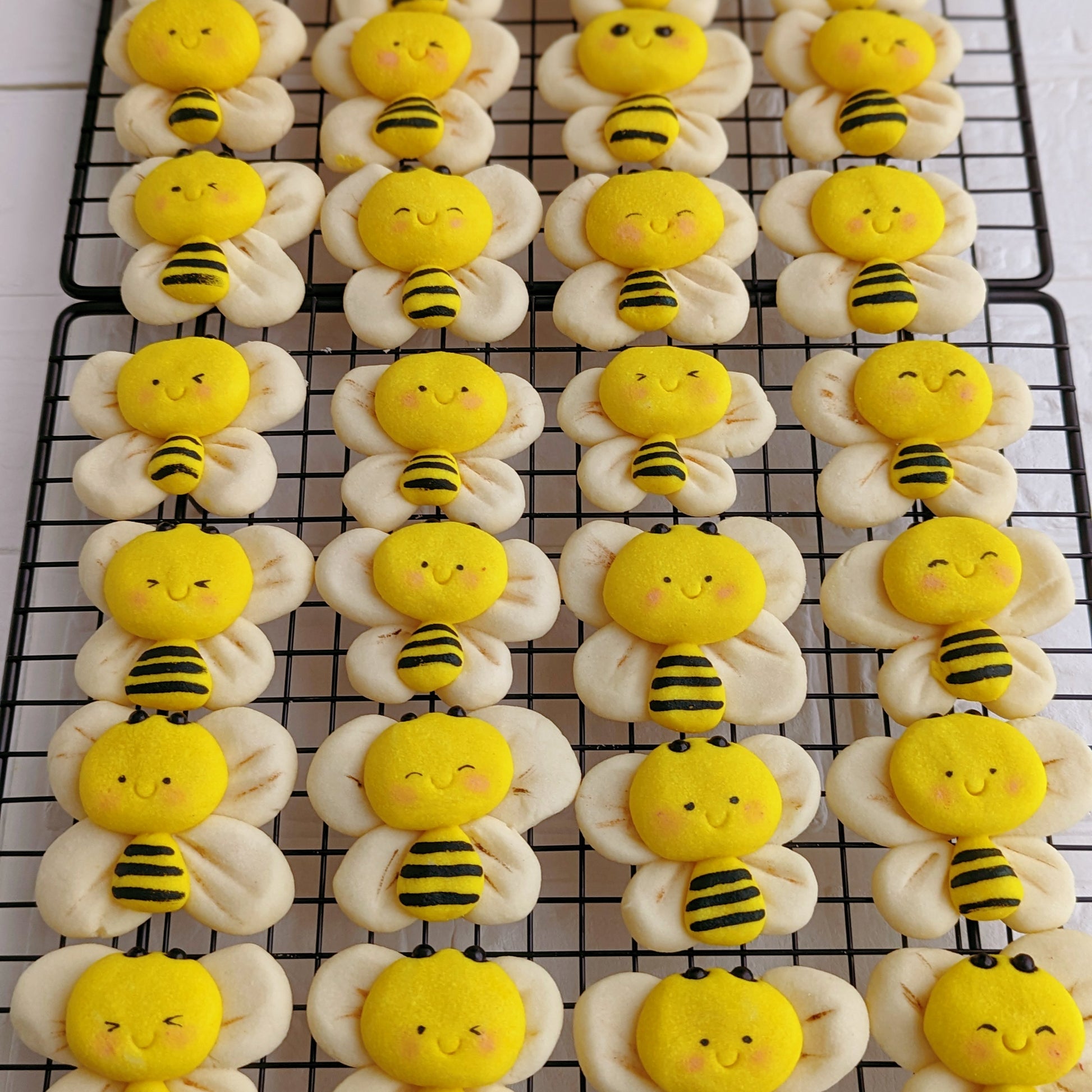 Rows of bumblebee-shaped sugar cookies cooling on a black wire rack. Each cookie has a pale cream-colored body and wings with a bright yellow face and striped black-and-yellow abdomen.