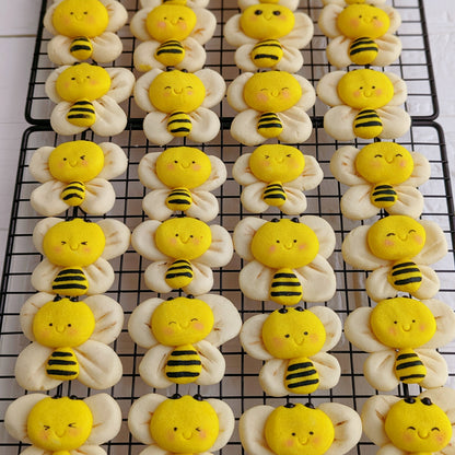 Rows of bumblebee-shaped sugar cookies cooling on a black wire rack. Each cookie has a pale cream-colored body and wings with a bright yellow face and striped black-and-yellow abdomen.
