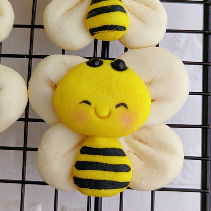 A close-up of two decorated baked cookies shaped like bumblebees with yellow bodies, black stripes, and cream-colored wings, arranged on a black wire cooling rack.