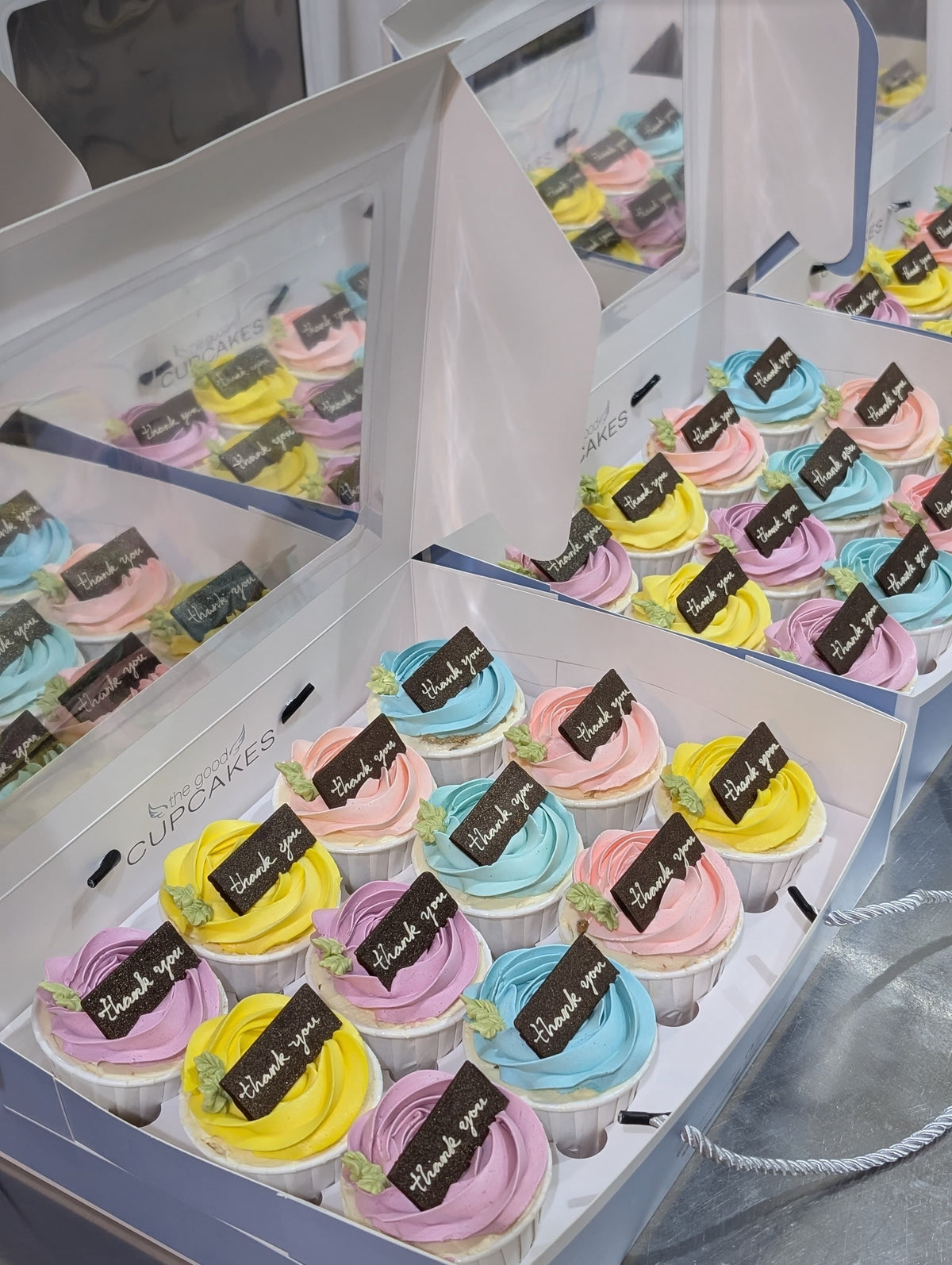 Open gift box containing twelve pastel pink, purple and aqua cupcakes with chocolate “thank you” plaques, surrounded by matching cupcake boxes.