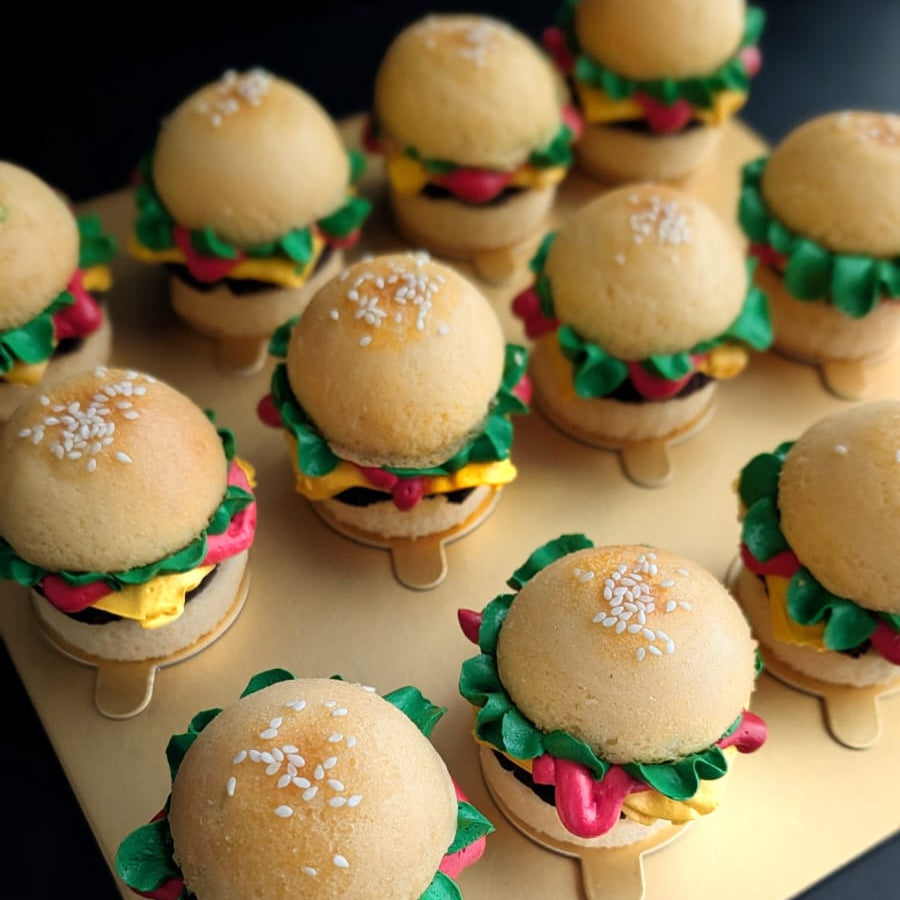 Box of 12 novelty burger cupcakes on a gold board — each built from two cupcake "buns" with a chocolate cake "patty," hand-piped green lettuce, yellow cheese and red tomato buttercream, and a sesame seed topping