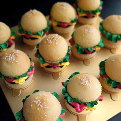 Box of 12 novelty burger cupcakes on a gold board — each built from two cupcake "buns" with a chocolate cake "patty," hand-piped green lettuce, yellow cheese and red tomato buttercream, and a sesame seed topping