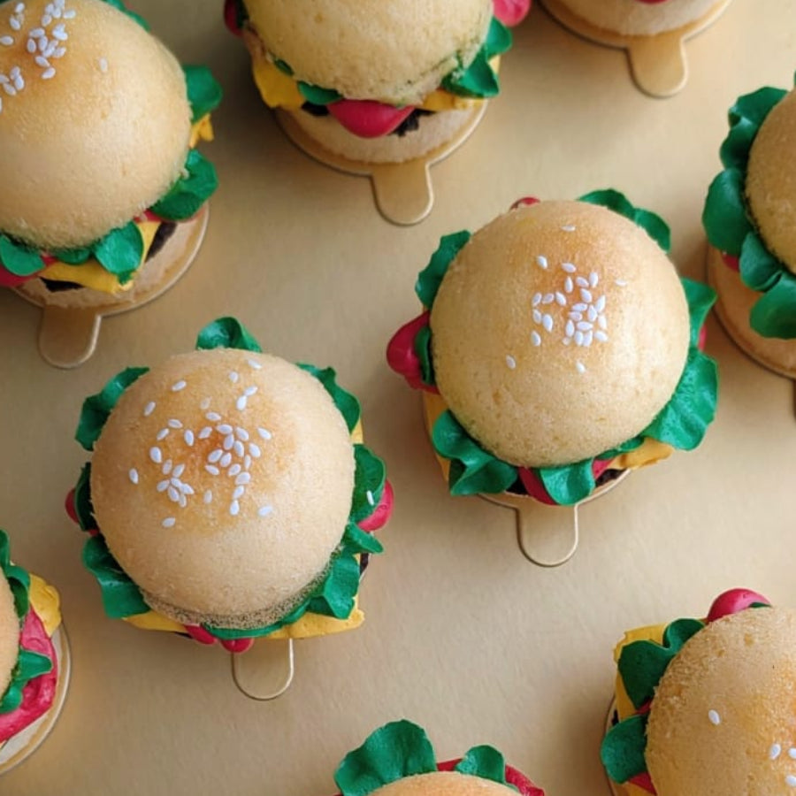 Box of 12 novelty burger cupcakes on a gold board — each built from two cupcake "buns" with a chocolate cake "patty," hand-piped green lettuce, yellow cheese and red tomato buttercream, and a sesame seed topping