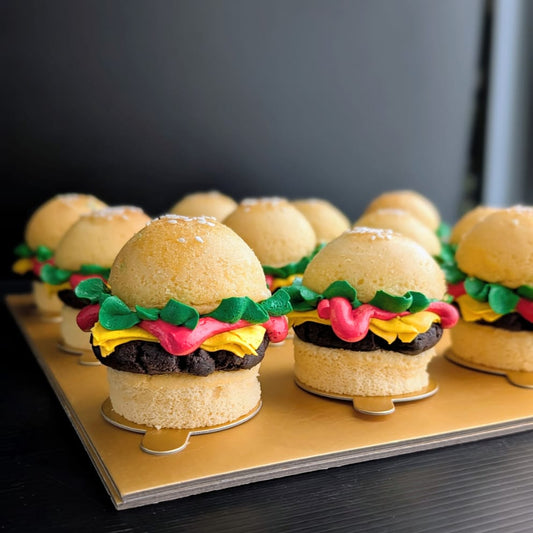 Box of 12 novelty burger cupcakes on a gold board — each built from two cupcake "buns" with a chocolate cake "patty," hand-piped green lettuce, yellow cheese and red tomato buttercream, and a sesame seed topping