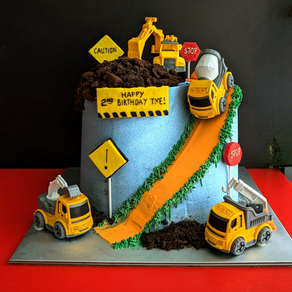Construction site birthday cake with toy trucks, excavator, cement mixer, chocolate dirt, orange road and green grass buttercream details, low-sugar cake Singapore