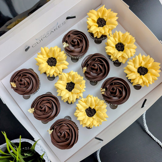 Box of 12 cupcakes — six dark chocolate rosette cupcakes with gold leaf accents and six hand-piped yellow sunflower cupcakes with a chocolate crumb centre, in a white gift box labelled The Good Cupcakes