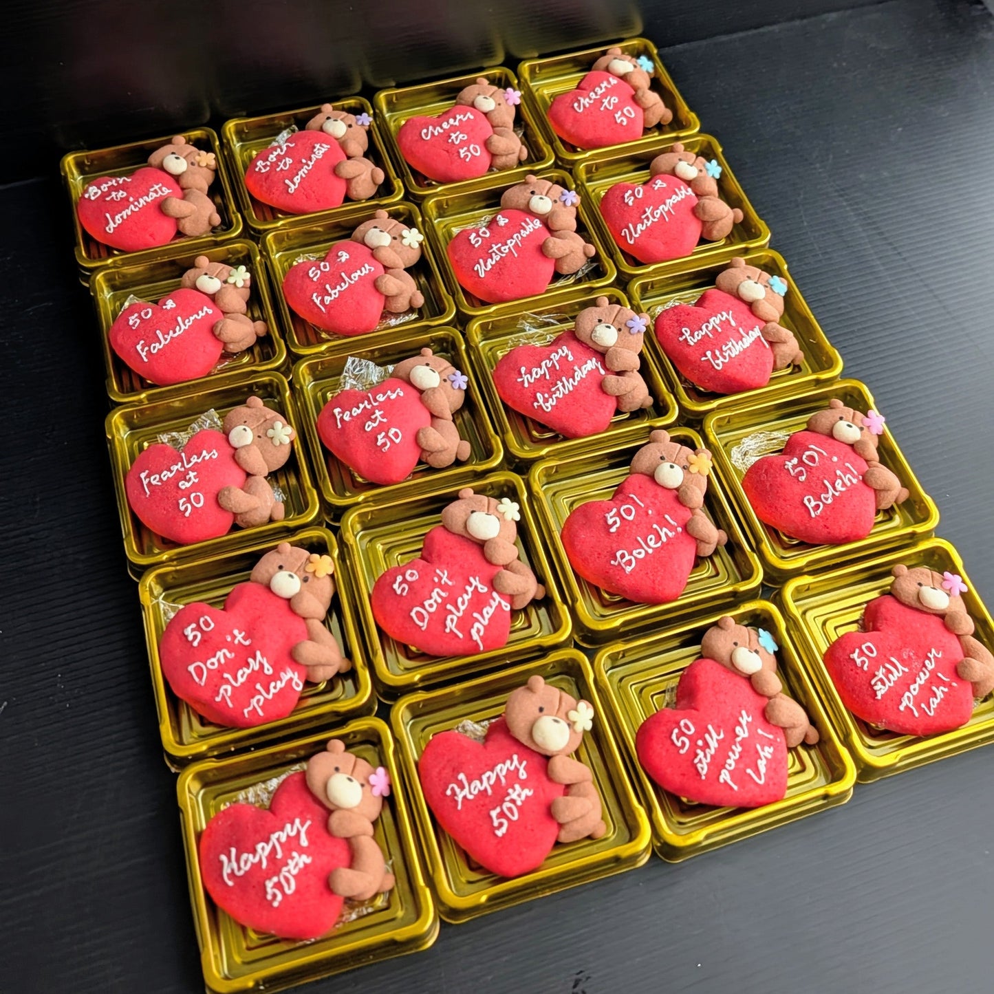 Custom handmade shortbread cookies individually boxed in gold gift boxes — red heart-shaped cookies with hand-painted teddy bear figures and personalised birthday messages including "Born to Dominate", "Cheers to 50", "50 & Fabulous", "Fearless at 50", and "Happy Birthday". Made to order by The Good Cupcakes, Singapore.