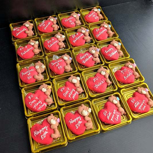 Custom handmade shortbread cookies individually boxed in gold gift boxes — red heart-shaped cookies with hand-painted teddy bear figures and personalised birthday messages including "Born to Dominate", "Cheers to 50", "50 & Fabulous", "Fearless at 50", and "Happy Birthday". Made to order by The Good Cupcakes, Singapore.