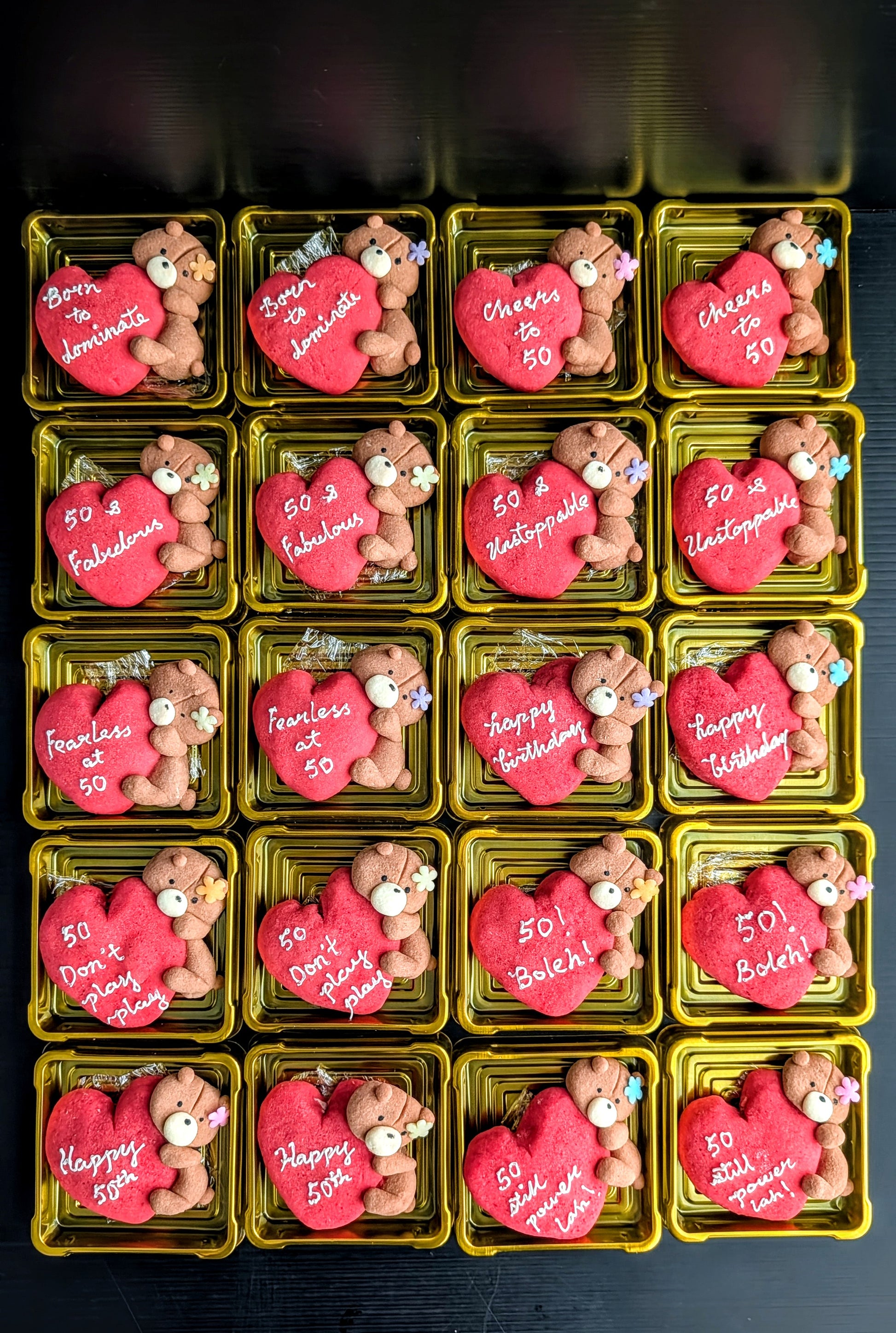 A grid of 16 small gold rectangular boxes, each containing a pink heart-shaped cookie decorated with a teddy bear and white Valentine's Day messages such as '50! Love Always,' 'Cheers to 50,' and 'Happy Valentine's Day.'.