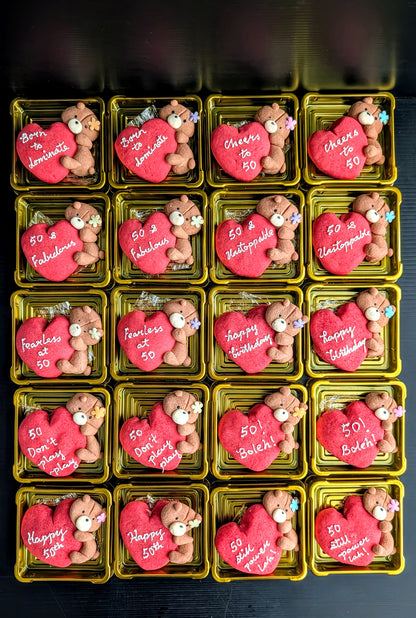 A grid of 16 small gold rectangular boxes, each containing a pink heart-shaped cookie decorated with a teddy bear and white Valentine's Day messages such as '50! Love Always,' 'Cheers to 50,' and 'Happy Valentine's Day.'.