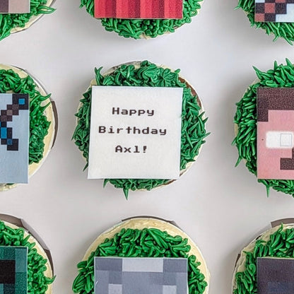 Box of cupcakes decorated with pixelated designs inspired by a popular video game, featuring characters and symbols.