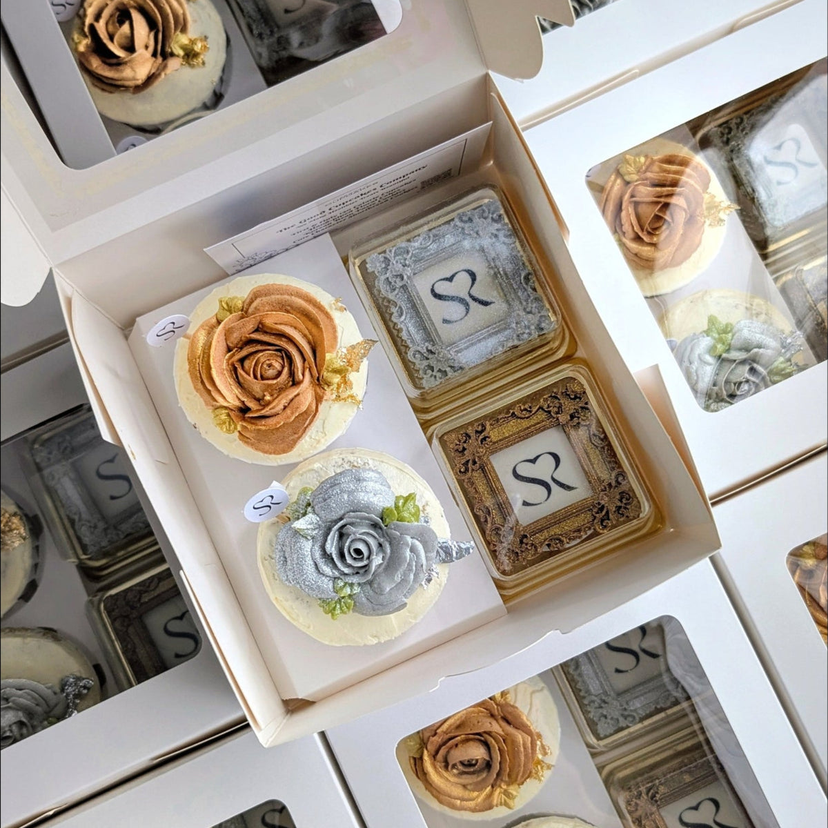 Open white gift box containing two metallic floral cupcakes and two ornate Sae-Ren Spa logo cookies, surrounded by matching boxes.