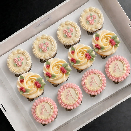 A boxed set of 12 vintage monogram cupcakes, from The Good Cupcakes, arranged in a white box  on a dark background. The cupcakes feature three styles: cream-colored roses with pink floral accents, cream roses with green leaves, and pink rosette-edged cupcakes with pale cream centers decorated with pink monogram letters 'A'.