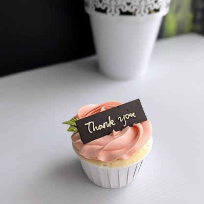 Close-up of thank you cupcake with pink rose frosting and chocolate message tag on white surface