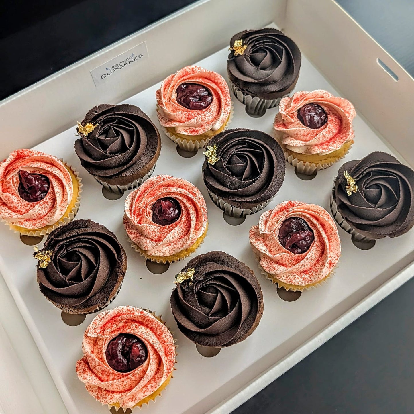 Box of 12 hand-piped rosette cupcakes — six dark chocolate Chocolate Chocolate cupcakes with gold leaf accents and six blush pink Strawberries & Cream cupcakes, in a white gift box labelled The Good Cupcakes