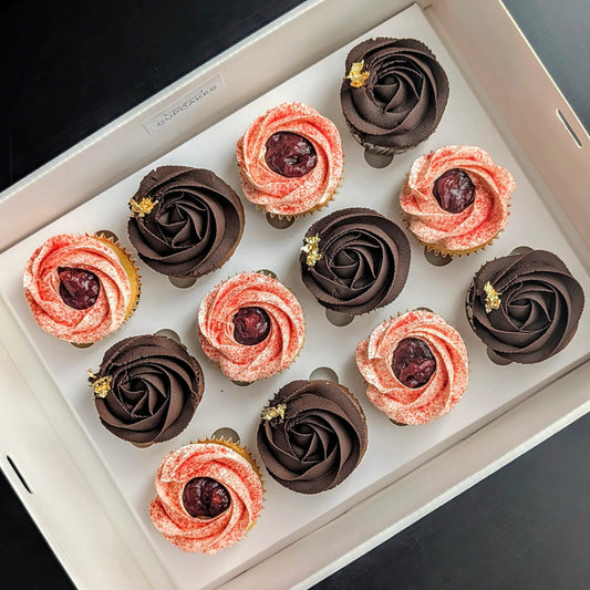 Box of 12 hand-piped rosette cupcakes — six dark chocolate Chocolate Chocolate cupcakes with gold leaf accents and six blush pink Strawberries & Cream cupcakes, in a white gift box labelled The Good Cupcakes