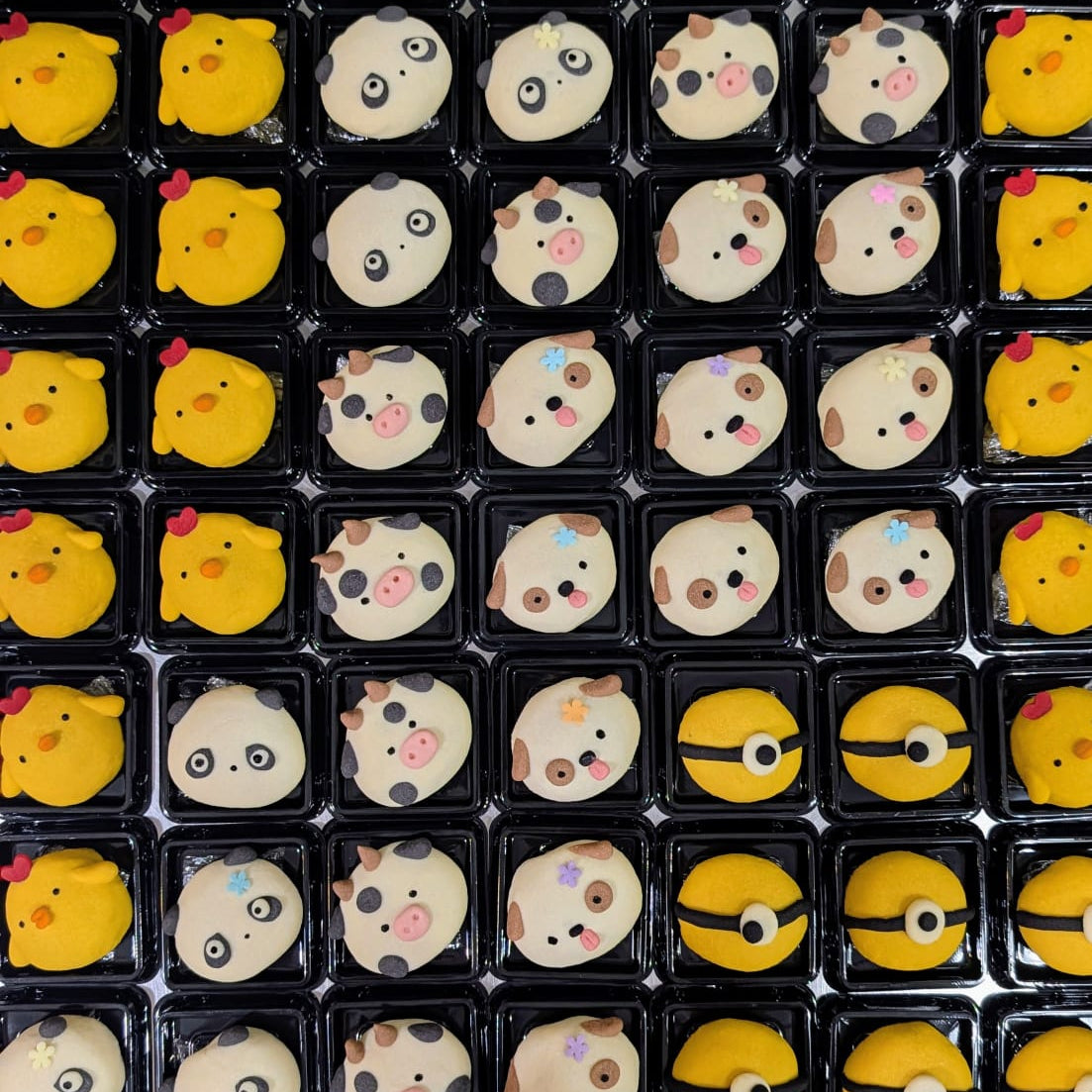 A grid of small black plastic trays holding round novelty cookies decorated with cartoon faces and animal motifs, including yellow chick-like faces, panda faces, cow faces, and a Pokéball face, arranged on a metallic surface.