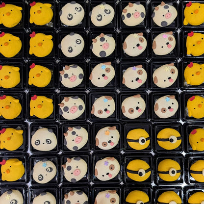 A grid of small black plastic trays holding round novelty cookies decorated with cartoon faces and animal motifs, including yellow chick-like faces, panda faces, cow faces, and a Pokéball face, arranged on a metallic surface.