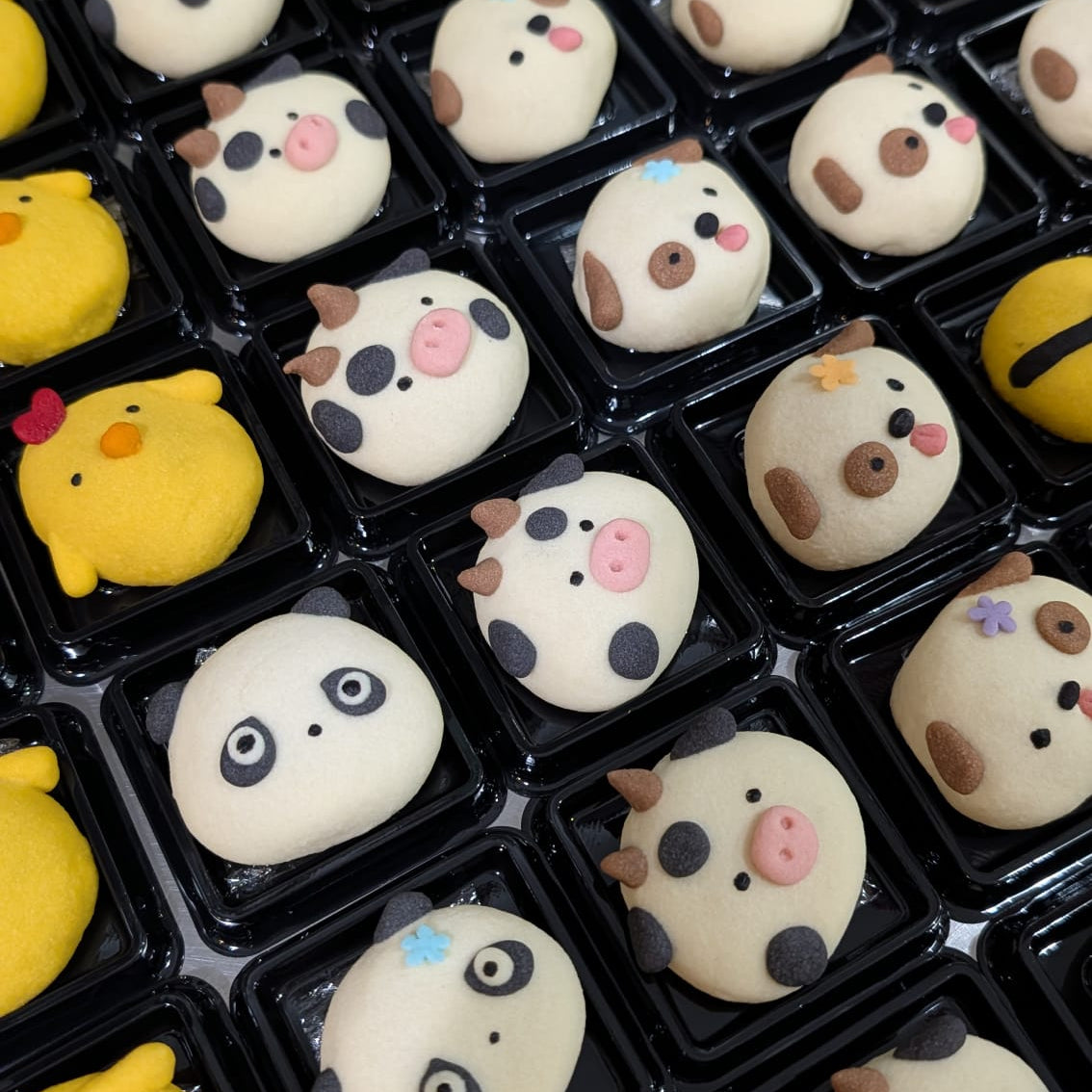 A close-up view of many small novelty cookies arranged in black plastic trays. The pastries are mostly off-white with cute cartoon faces in black, brown, pink, and blue, plus several bright yellow chick-shaped cookies.
