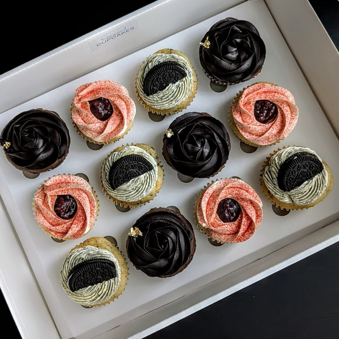 A box of assorted cupcakes arranged in a white tray against a dark background. The cupcakes include dark chocolate, pink, and light beige swirled frosting with dark berry centers.