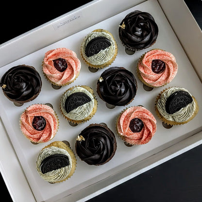 A box of assorted cupcakes arranged in a white tray against a dark background. The cupcakes include dark chocolate, pink, and light beige swirled frosting with dark berry centers.