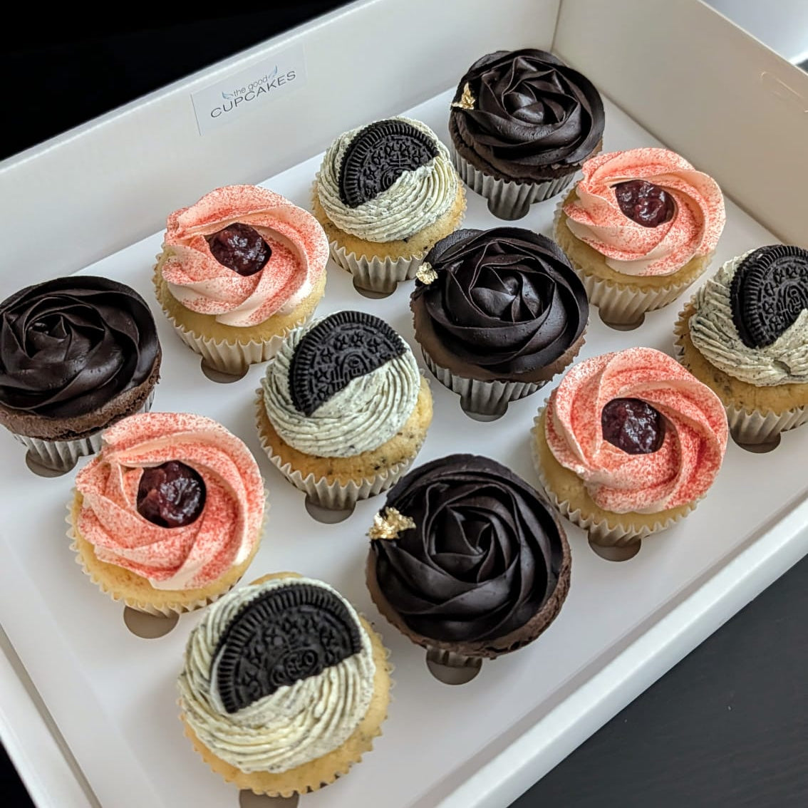 A white box containing 12 assorted cupcakes with various frosting designs including pink, chocolate, and cream swirls, some topped with dark berries or Oreo cookies, set against a dark background.