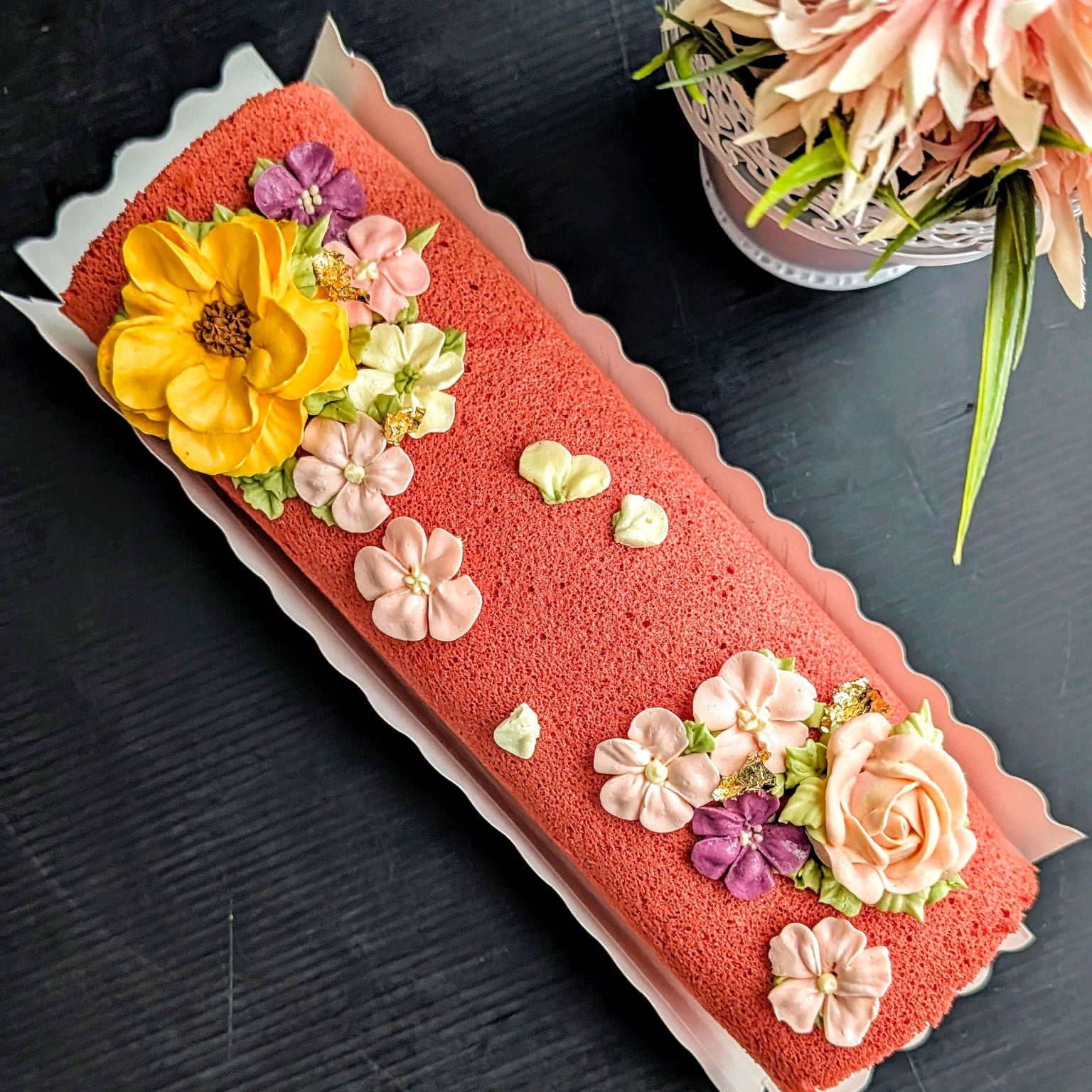 A long roll cake with a coral-red sponge base, decorated with large yellow, pink, peach, and purple flower decorations, photographed on a dark tabletop with a floral arrangement in the corner.