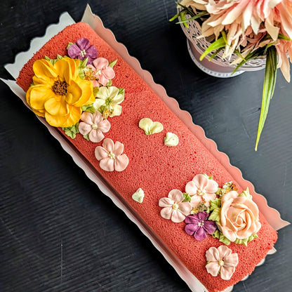 A long roll cake with a coral-red sponge base, decorated with large yellow, pink, peach, and purple flower decorations, photographed on a dark tabletop with a floral arrangement in the corner.