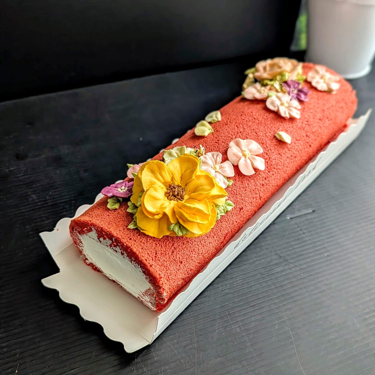 A red velvet cake roll decorated with a large yellow flower, smaller pink and yellow flowers, and green leaves, served on a white paper liner on a dark wooden table.